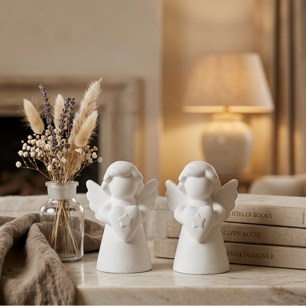 Dois mini anjos de cerâmica perfumada branca junto a uma jarra com flores secas e livros.