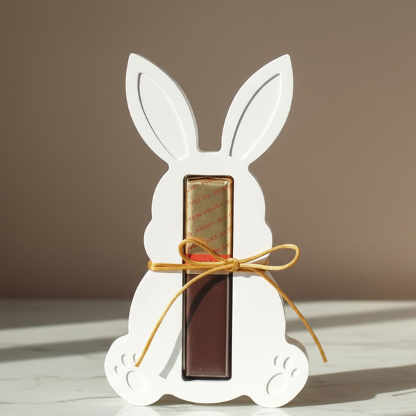 Coelho decorativo branco com laço dourado segurando bombom de chocolate para presente de Páscoa.