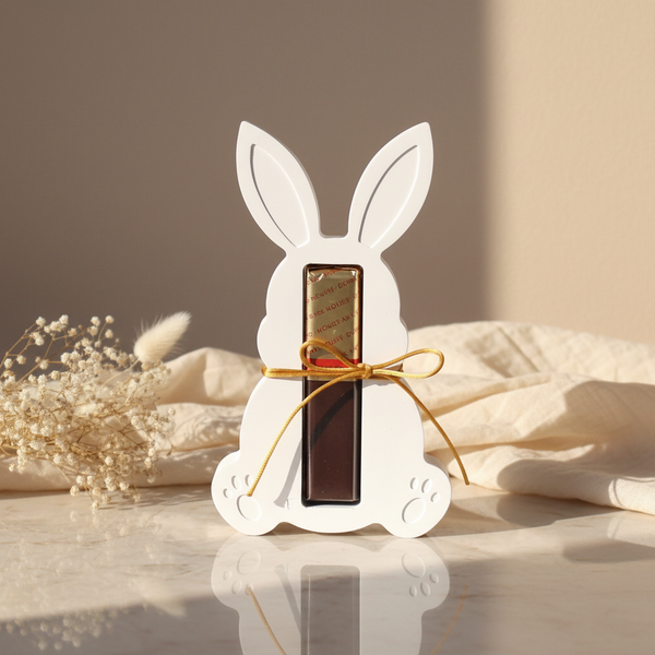 Coelho decorativo branco com laço dourado segurando bombom de chocolate para presente de Páscoa.