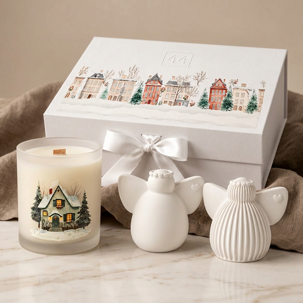 Dois anjos brancos de cerâmica perfumada, vela com ilustração de casa natalícia e caixa de oferta, junto a tecido bege.