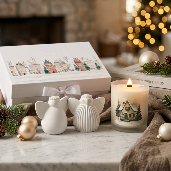 Vela acesa junto a dois anjos de cerâmica perfumada e caixa de oferta natalícia, com luzes de Natal ao fundo