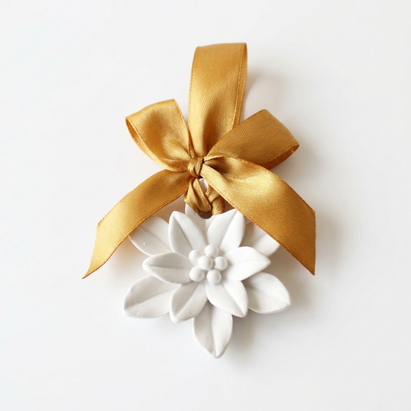 Flor Estrela de Natal de cerâmica perfumada branca, com fita e laço dourados, sobre fundo claro.