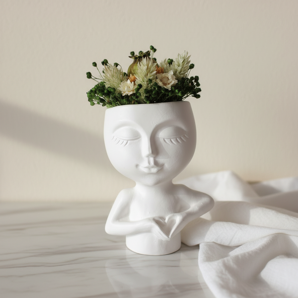 Jarra decorativa rosto feminino com mãos formando coração e arranjo de flores secas.