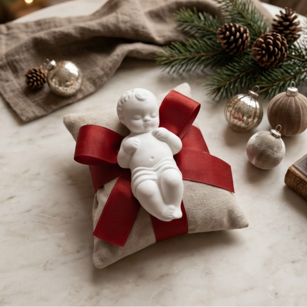 Menino Jesus de cerâmica perfumada branca sobre almofada bege com laço vermelho, numa composição de Natal.