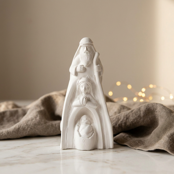 Sagrada Família Alta branca sobre mármore, com tecido bege e luzes desfocadas ao fundo.