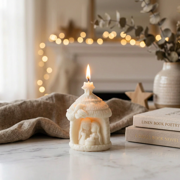 Vela Presépio acesa sobre mármore, junto a livros e tecido bege, com luzes de Natal ao fundo.