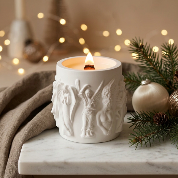 Vela de Natal acesa em pote de cerâmica branca com anjos em relevo, junto a ramos e bolas de Natal.